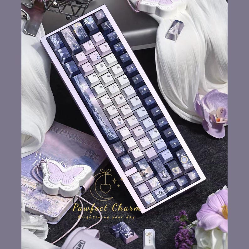 May include: A custom mechanical keyboard with a lavender and white color scheme. The keycaps feature a gradient design and various decorative icons. The keyboard is surrounded by accessories, including a butterfly-shaped item and a book.