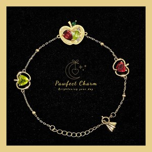May include: A gold-toned chain bracelet featuring three apple-shaped charms with red and green heart-shaped stones. The central charm includes a small green leaf and clear accents. The text "Pawfect Charm Brightening your day" is centered.