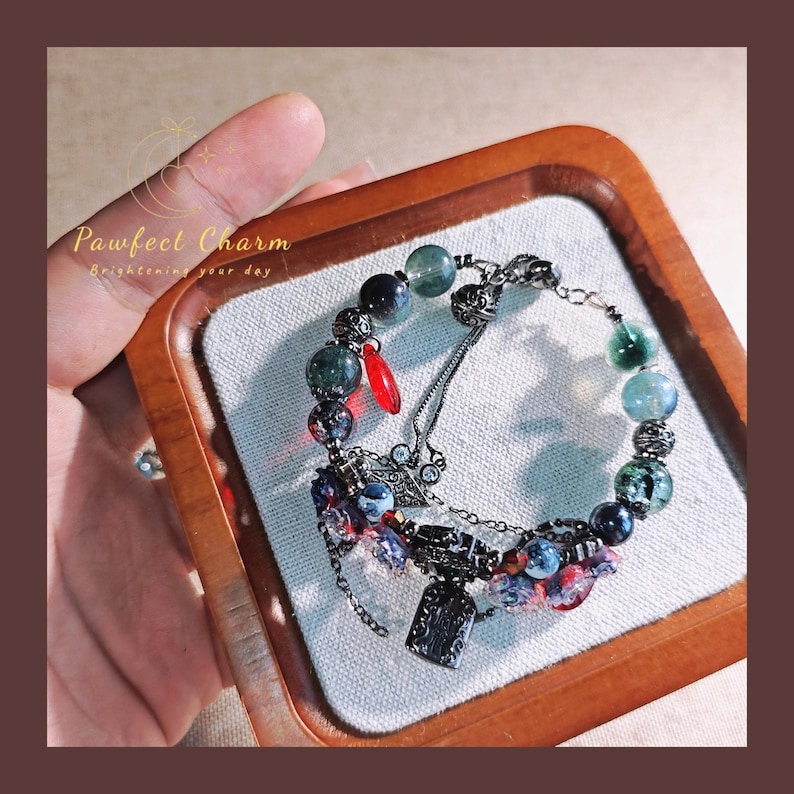 May include: A beaded bracelet with a mix of dark blue, green, and red beads. The bracelet includes a small red charm and a decorative metal tag. The text "Pawfect Charm Brightening your day" is visible.