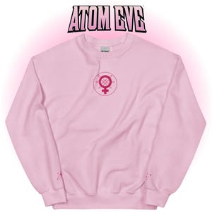 May include: A light pink sweatshirt with the text "ATOM EVE" in a bold, outlined font at the top. The sweatshirt features a pink graphic on the front and a small design on the sleeve.