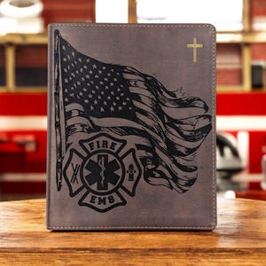 May include: A brown leather-like journal featuring an engraved American flag and the Fire EMS emblem. The flag is waving, and the emblem includes a Maltese cross, a snake, and a fire hydrant. The journal has a stitched border.