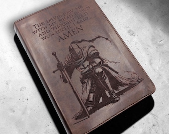 Könnte beinhalten: Braunes Lederjournal mit einem geprägten Bild eines knienden Ritters mit einem Schwert. Der Einband des Journals trägt den Text "THE DEVIL SAW ME WITH MY HEAD DOWN AND THOUGHT HE'D WON UNTIL I SAID... AMEN."