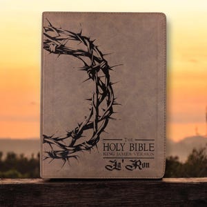 Może przedstawiać: Jasnobrązowa skórzana Biblia z czarną ilustracją korony cierniowej. Na okładce widnieje napis "THE HOLY BIBLE KING JAMES VERSION" oraz imię "Ja' Ron". Tło przedstawia zachód słońca.