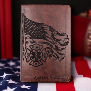 May include: Brown leather journal featuring an engraved American flag and a fire department emblem. The text "FIRE EMS" and the name "Austin Harrold" are also engraved on the cover. The journal is a patriotic item.