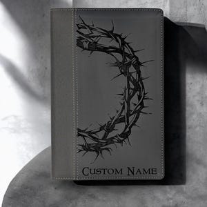 Op de afbeelding: Grijs lederen dagboek met een zwart doornenkroonontwerp en de tekst "CUSTOM NAME" op de voorkant.