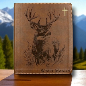May include: Brown leather journal with a detailed engraved image of a buck deer. The journal features the name "Wyatt Martin" at the bottom. The background shows a mountain landscape.