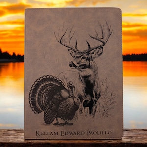 May include: A tan leather journal featuring a detailed illustration of a buck and a turkey. The buck has large antlers, and the turkey has its feathers fanned out. The text "KELLAM EDWARD PAOLILLO" is printed at the bottom.