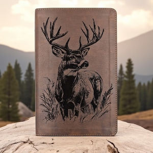 May include: A brown leather-like tablet case featuring a detailed black illustration of a buck deer with large antlers. The deer is depicted in a grassy field, with a mountain and forest backdrop. The case has a textured surface.