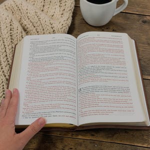 May include: Open Bible with brown leather cover, pages with text, and a white coffee mug in the background. A hand is visible, holding the book open. The text is in English.