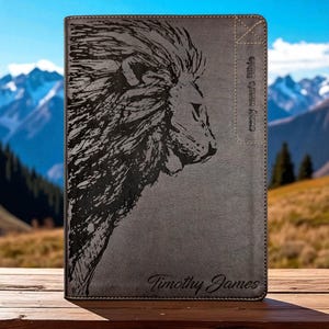 Puede incluir: Cubierta de Biblia gris con una detallada ilustración en negro de la cabeza de un león. La cubierta tiene el texto "every man's bible" y "Timothy James". El fondo muestra una cordillera.