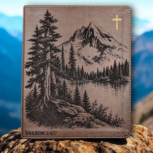 May include: A brown leather-bound journal with a mountain and lake landscape design. The cover features a detailed engraving of a forest, lake, and mountain range. A gold cross is in the upper right corner, and the word "Valencia" is at the bottom.