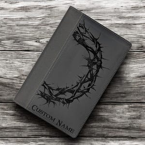 Op de afbeelding: Een grijs lederen dagboek met een zwart doornenkroonontwerp en de tekst "CUSTOM NAME" gegraveerd op de voorkant.