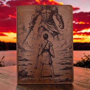 May include: A rectangular tan leather-like journal cover featuring a laser-engraved illustration of a small figure facing a giant armored warrior holding a sword. The name JOSHUA RYAN WOODSON is engraved in a serif font at the bottom of the cover.