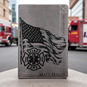 May include: Gray Bible cover featuring an American flag and a Fire EMS emblem. The text "every men's Bible" is on the side, and "Matt Hogan" is at the bottom. The cover is made of a textured material.