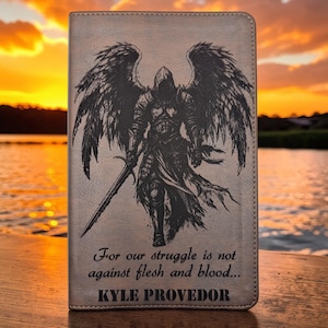 Könnte beinhalten: Ein rechteckiges Notizbuch mit hellbraunem Einband und einer schwarzen Lasergravur eines geflügelten Kriegers mit Schwert. Der Text lautet: For our struggle is not against flesh and blood... sowie der Name KYLE PROVEDOR.