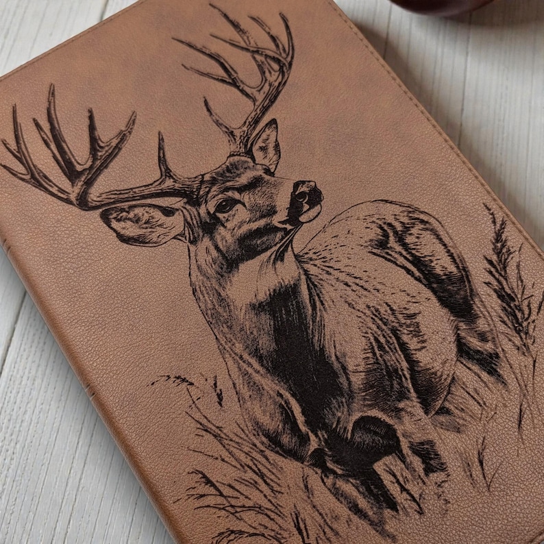 May include: A brown leather journal with a detailed engraved image of a buck deer with large antlers. The deer is depicted in a naturalistic style, surrounded by tall grass. The journal has a textured surface.