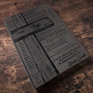 May include: A rectangular gray leather-like journal cover featuring a dark engraved wooden cross graphic and various religious scripture verses. The text includes phrases like For we live by faith, not by sight and the name Bruce Smith is engraved.