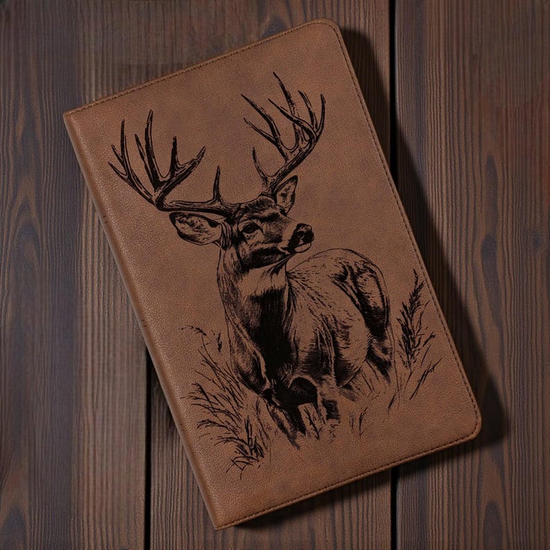 May include: A tan leather-bound journal featuring a detailed black engraving of a buck deer with large antlers. The deer is depicted in a grassy field. The journal is rectangular and has rounded corners.