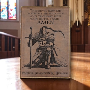 Könnte beinhalten: Ein braunes, ledergebundenes Buch mit goldenem Rand. Auf dem Einband ist ein kniender Ritter mit Schwert abgebildet, sowie der Text "THE DEVIL SAW ME WITH MY HEAD DOWN AND THOUGHT HE'D WON UNTIL I SAID... AMEN." Der Name des Autors ist ebenfalls aufgedruckt.