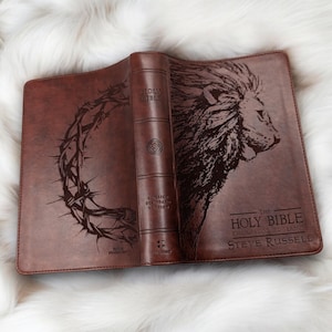 May include: Open brown leather Bible with a lion's head and a crown of thorns design. The text "The Holy Bible" and "English Standard Version" are visible. The book is open on a white fluffy surface.
