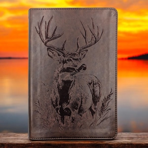 May include: A brown leather journal with a detailed engraved image of a buck deer with large antlers. The deer is depicted in a grassy field. The background shows a sunset over a lake.