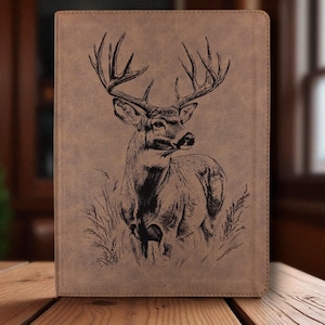May include: A brown leather-like journal cover with a detailed black engraving of a buck deer in a grassy field. The deer has large antlers and is facing to the right. The cover has a rectangular shape.