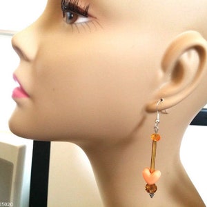 Puede incluir: Un pendiente colgante con un gancho plateado. El pendiente presenta una cuenta en forma de coraz&oacute;n de color melocot&oacute;n, cuentas de color &aacute;mbar y una varilla larga y delgada de color &aacute;mbar. El pendiente se muestra en un maniqu&iacute;.