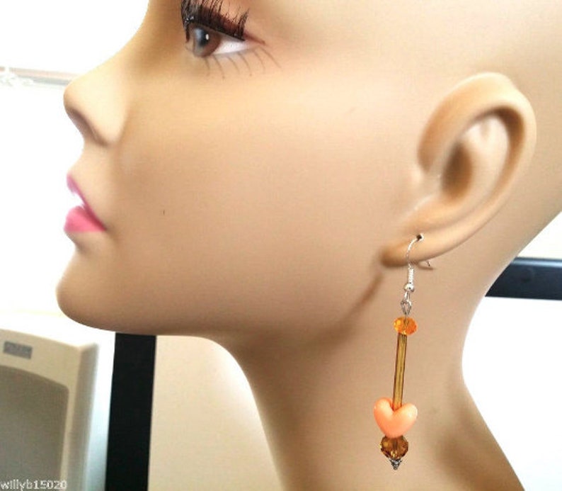 Puede incluir: Un pendiente colgante con un gancho plateado. El pendiente presenta una cuenta en forma de coraz&oacute;n de color melocot&oacute;n, cuentas de color &aacute;mbar y una varilla larga y delgada de color &aacute;mbar. El pendiente se muestra en un maniqu&iacute;.