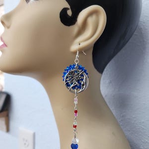 May include: Silver-tone dangle earrings featuring a celestial design. Each earring has a blue beaded circle with a silver crescent moon and a figure. A strand of colorful beads hangs below, ending with a blue and clear bead.
