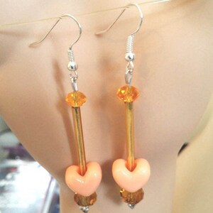 Puede incluir: Un par de pendientes colgantes con ganchos plateados. Cada pendiente presenta una cuenta en forma de coraz&oacute;n melocot&oacute;n, un tubo dorado y cuentas de cristal naranja y marr&oacute;n. Los pendientes est&aacute;n dise&ntilde;ados para colgar de la oreja.