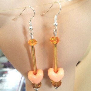 Puede incluir: Un par de pendientes colgantes con ganchos plateados. Cada pendiente presenta una cuenta en forma de coraz&oacute;n de color melocot&oacute;n, un tubo dorado y cuentas de cristal de color naranja y &aacute;mbar. Los pendientes est&aacute;n dise&ntilde;ados para colgar de la oreja.