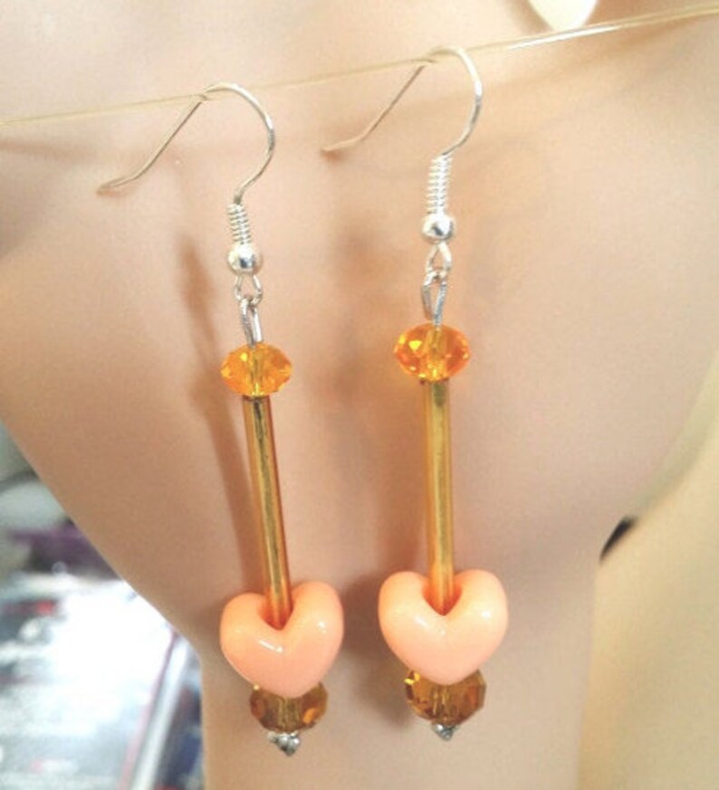 Puede incluir: Un par de pendientes colgantes con ganchos plateados. Cada pendiente presenta una cuenta en forma de coraz&oacute;n de color melocot&oacute;n, un tubo dorado y cuentas de cristal de color naranja y &aacute;mbar. Los pendientes est&aacute;n dise&ntilde;ados para colgar de la oreja.