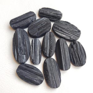 Può includere: Una collezione di undici pietre di tormalina nera levigate, ognuna caratterizzata da una distinta forma ovale allungata con striature verticali visibili e una consistenza superficiale liscia e riflettente sul materiale minerale scuro.