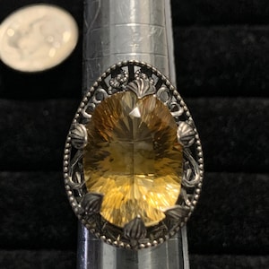 May include: A silver ring with an oval, golden-yellow gemstone. The ring has an ornate, filigree design around the stone, with decorative elements. A coin is visible in the background.