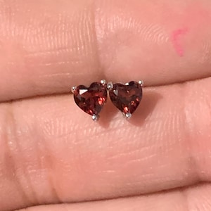 May include: A pair of heart-shaped stud earrings featuring deep red faceted gemstones set in silver-toned metal prongs. Each earring displays a clear, dark red hue with a polished finish, showcasing a classic heart silhouette and secure metal settings.