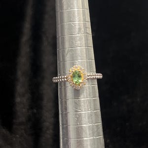 May include: A silver ring with a gold-colored halo around a light green oval gemstone. The halo is surrounded by small, clear gemstones. The ring is on a silver ring sizer.