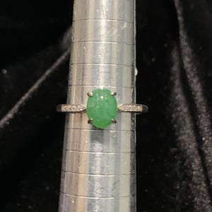 May include: A silver ring with a bright green oval gemstone at the center. The band is adorned with small, clear stones on either side of the central gem. The ring is displayed on a silver ring sizer against a dark background.