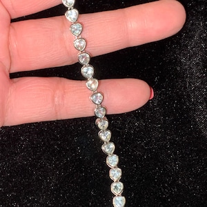 May include: A silver bracelet featuring heart-shaped clear gemstones. The bracelet is held against a dark background, showcasing the sparkling stones. The hearts are evenly spaced and linked together, creating a delicate and elegant piece of jewelry.