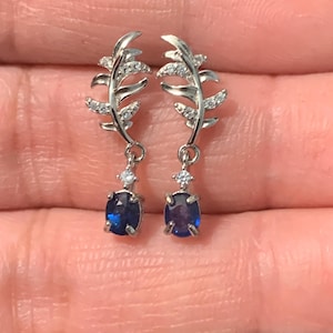 May include: A pair of silver-toned leaf-shaped earrings. Each earring features a delicate leaf design with small, clear stones and a dangling, oval-shaped, deep blue gemstone. The earrings are designed to be worn on the ear.