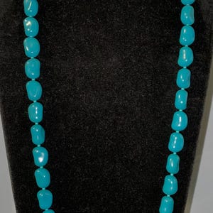 May include: A long necklace featuring turquoise-colored, irregular-shaped beads. The beads are strung together, creating a continuous loop. The necklace is displayed on a black mannequin.