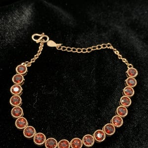 May include: A gold-toned bracelet featuring a series of round, red gemstones. The gemstones are set in a beaded bezel design, and the bracelet has a delicate chain with a lobster clasp. The jewelry is displayed on a dark background.