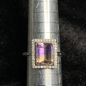 May include: A silver ring with a large, rectangular, purple and yellow gemstone surrounded by a halo of small, clear diamonds.