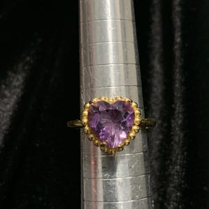 May include: A gold-toned ring with a heart-shaped, faceted purple gemstone. The gemstone is surrounded by a beaded gold border. The ring is displayed on a silver ring sizer against a dark background.