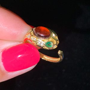 May include: A gold-toned open ring featuring a central oval-shaped dark red stone, accented with smaller green teardrop-shaped stones and tiny clear white stones embedded along the textured, snake-like metal band design.