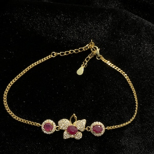May include: A gold-colored bracelet with a delicate chain. The bracelet features a floral design with a central ruby and surrounding clear stones. Two additional ruby-colored stones are set on either side of the floral design.