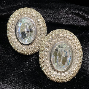 May include: A pair of oval-shaped earrings featuring a large, clear, faceted central stone surrounded by a dense border of small, shimmering white crystals set in a silver-toned metal frame, creating a textured and reflective appearance.