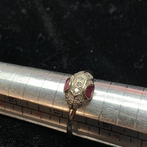 May include: A silver ring with a red and white stone setting. The ring is on a ruler, and the stone setting is in the shape of a flower.