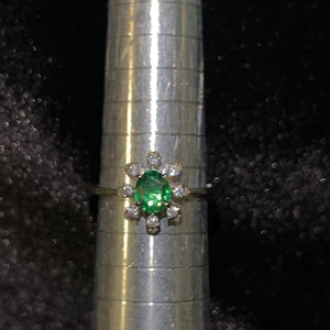 May include: A silver-toned ring featuring a central oval-cut green gemstone surrounded by eight small, clear, round-cut stones in a floral arrangement, positioned on a metal ring mandrel with visible horizontal measurement markings.