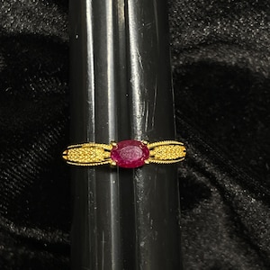 May include: A gold ring with a red oval gemstone set in the center. The band is decorated with a delicate, textured pattern.
