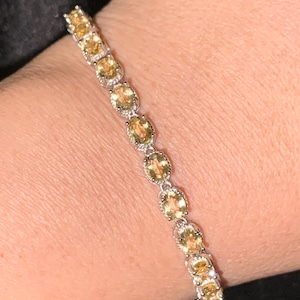 May include: A silver bracelet with oval-shaped, yellow gemstones. The bracelet is worn on a wrist, and the gemstones are set in a row. The bracelet has a delicate and elegant design.